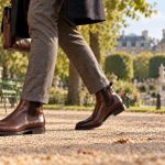 Homme marchant sur une allée gravillonnée en bottines pour homme britanniques marron et pantalon à carreaux, avec un sac en cuir et un parc en arrière-plan
