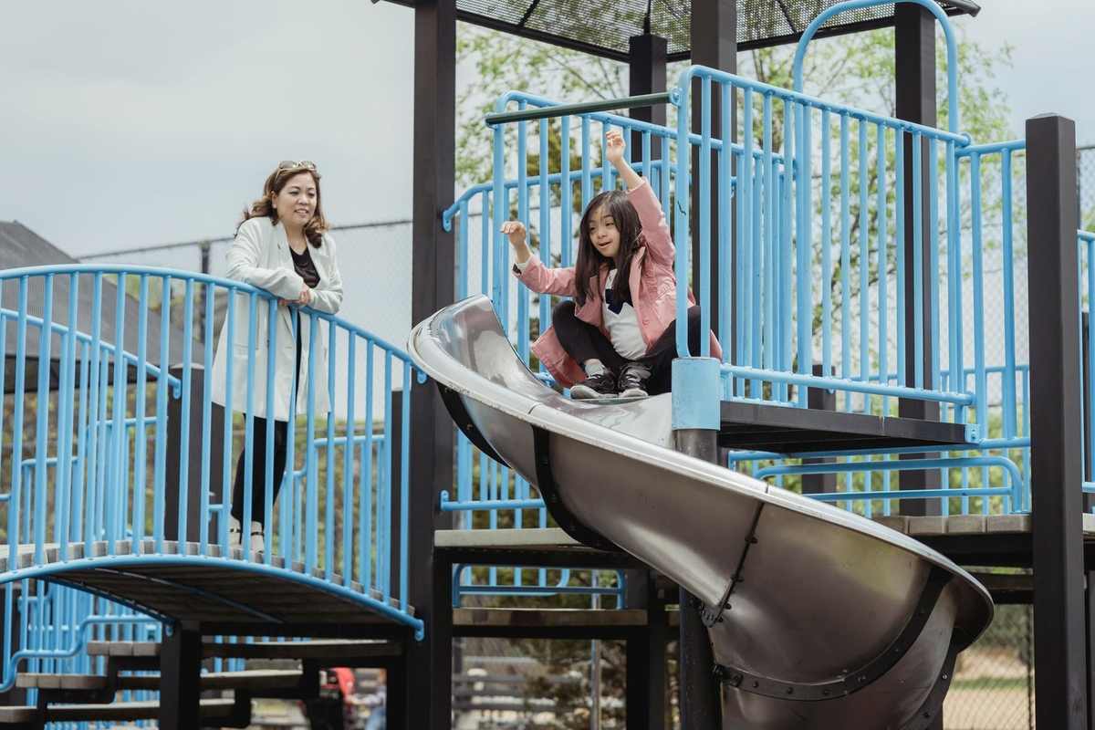 Enfant descend un toboggan en métal, adulte observe depuis une plateforme, structure de jeu avec barrières bleues Enfant descend un toboggan en métal, adulte observe depuis une plateforme, structure de jeu avec barrières bleues